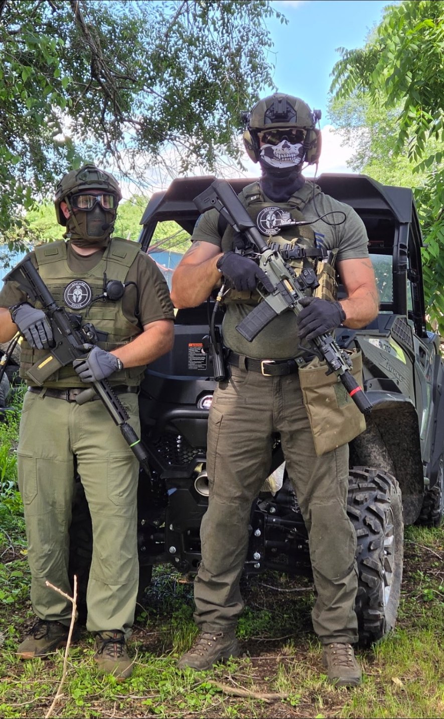 Two players in tactical vests, helmets and face protection hold airsoft rifles while posing in front of an off-road utility vehicle