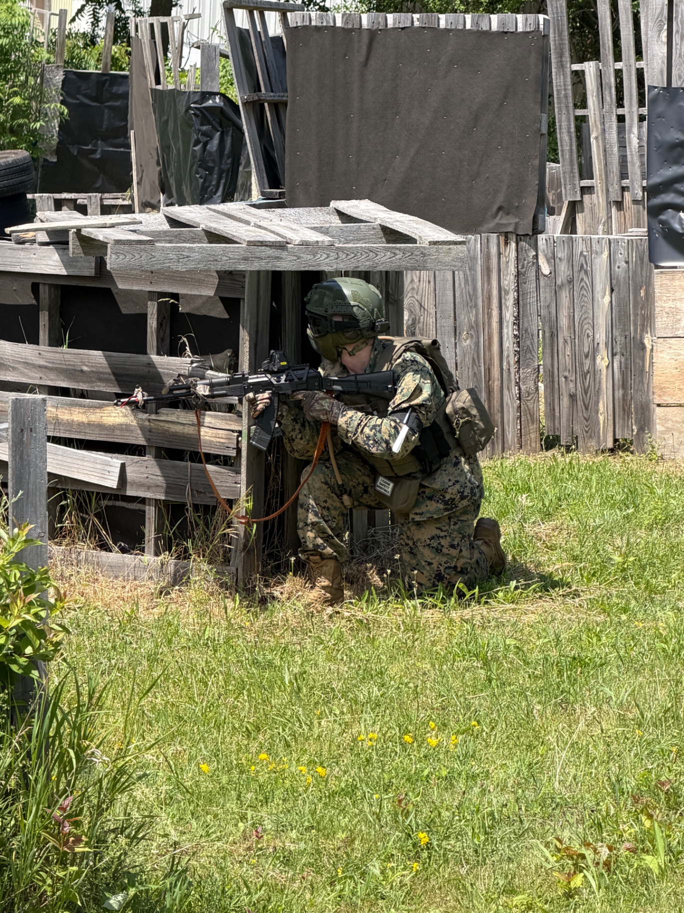 A player in digital camouflage, vest, helmet and protective mask kneels and aims a rifle from behind a wooden pallet bunker