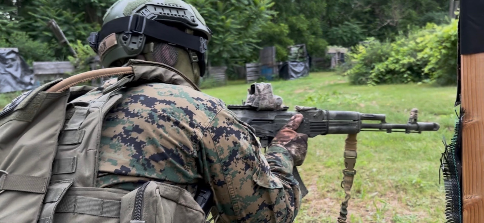 A player seen from behind in digital camouflage, helmet and plate carrier points an AK-pattern rifle toward an outdoor course of grass and trees
