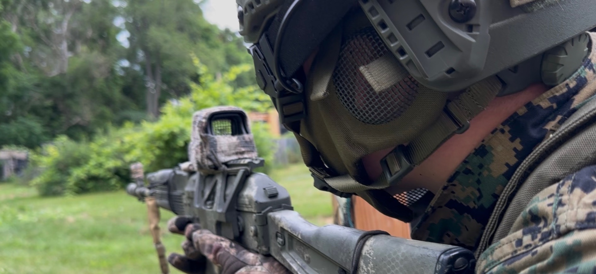 A player in an olive helmet with mesh face protection and digital camouflage aims an AK-style rifle fitted with a camouflaged optic across a grassy field