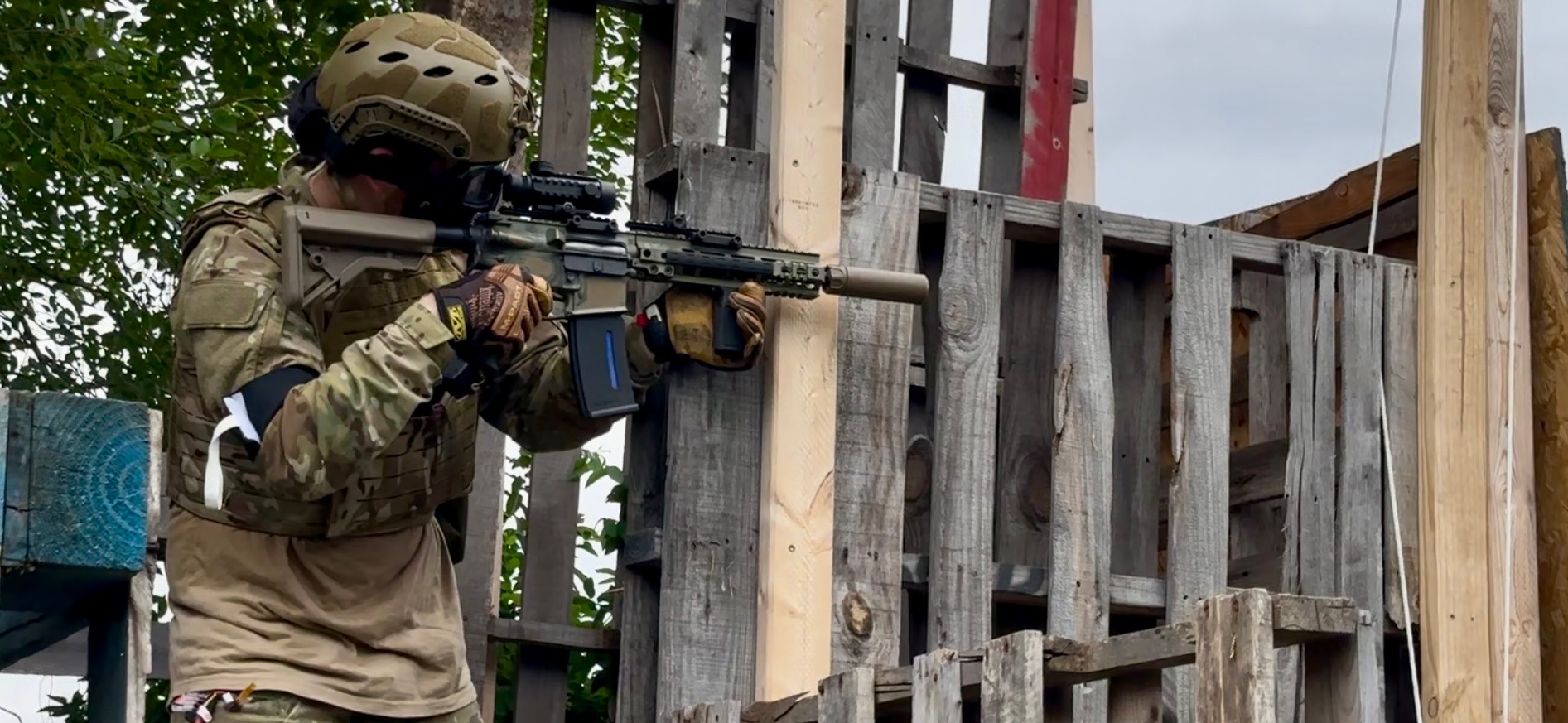 A player in full camouflage, helmet and eye protection aims a rifle through a pallet-built structure on the field