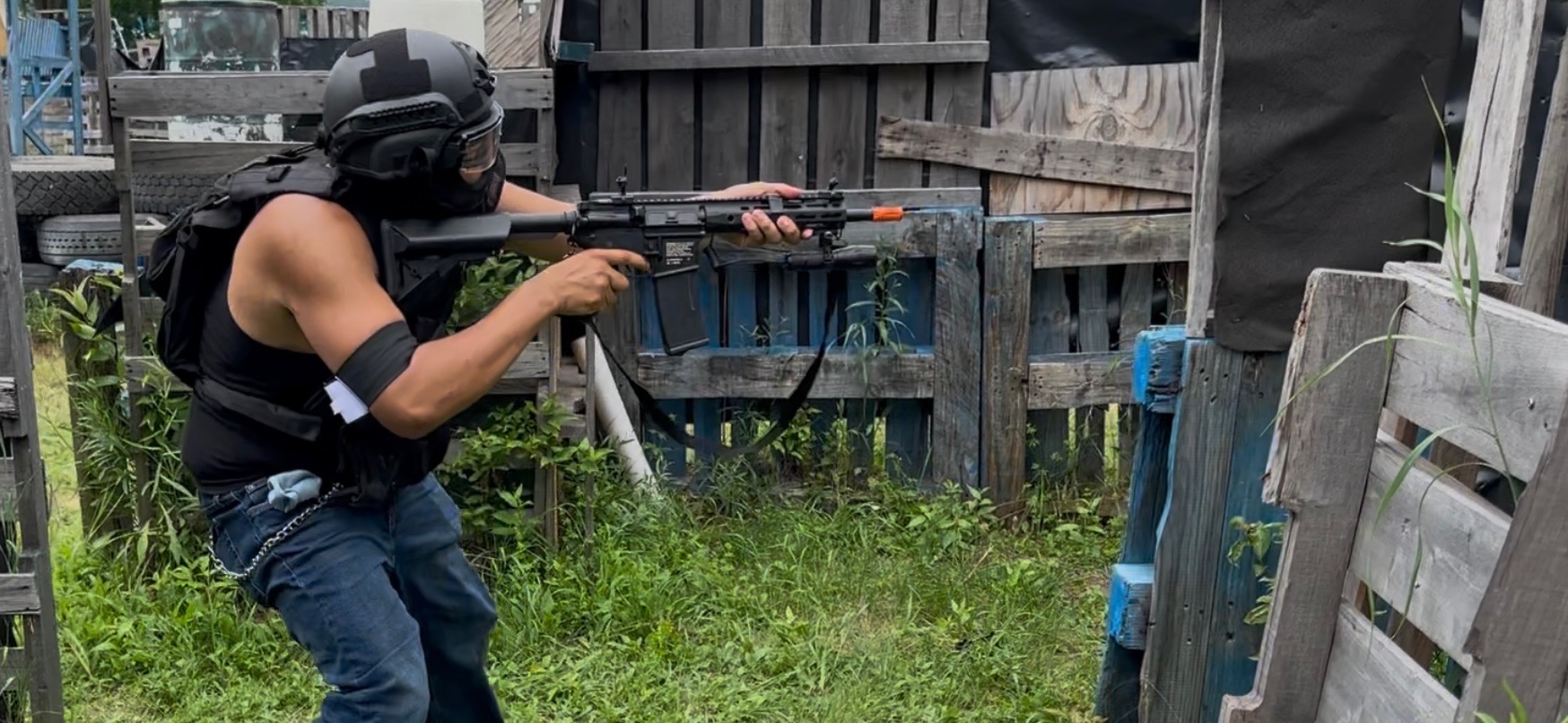 A player in a black helmet, goggles and tactical vest aims an orange-tipped airsoft rifle around wooden pallet obstacles
