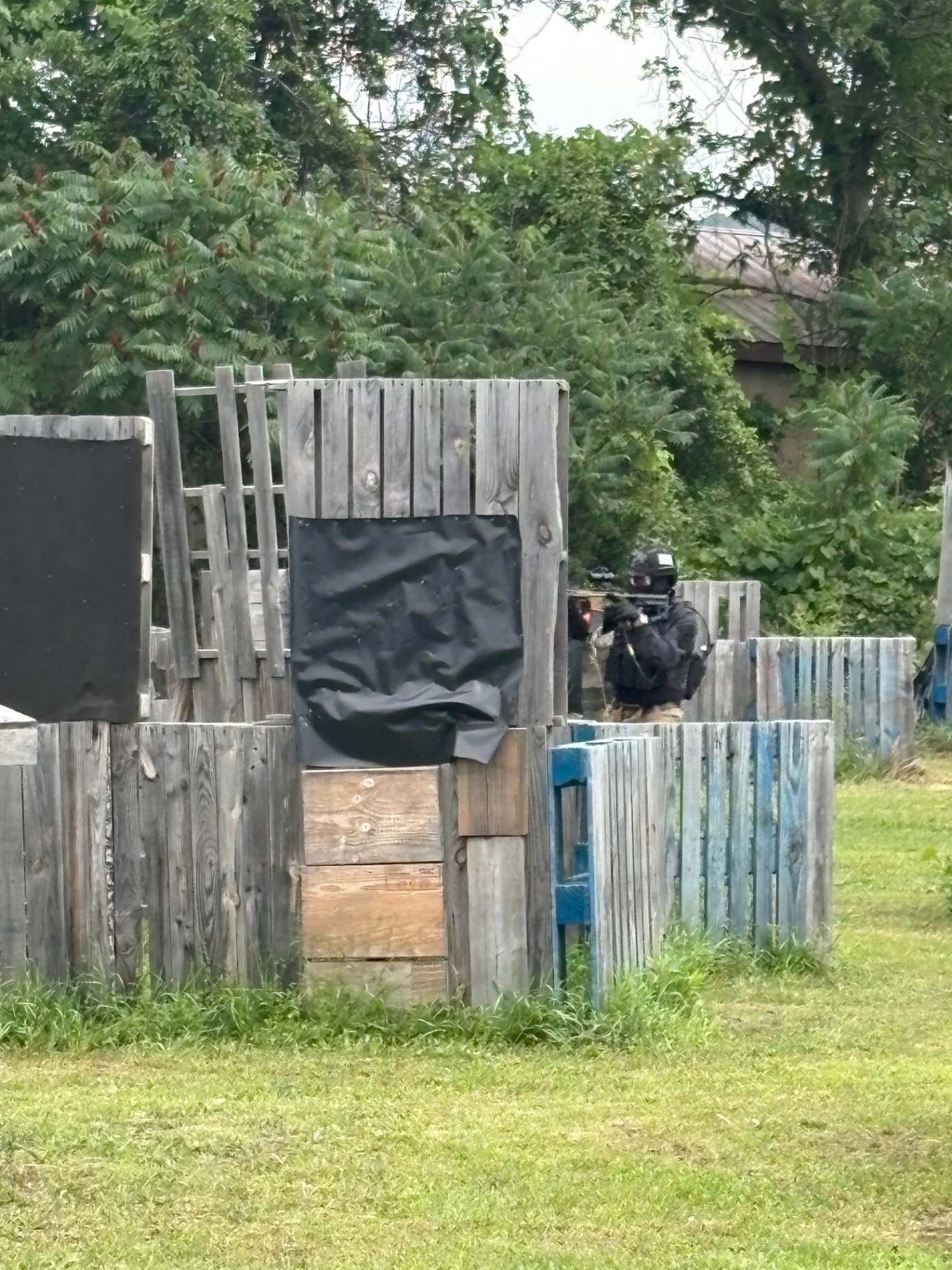 A player in black tactical gear, helmet and mask aims a rifle from behind a pallet barricade fitted with black tarp sheeting