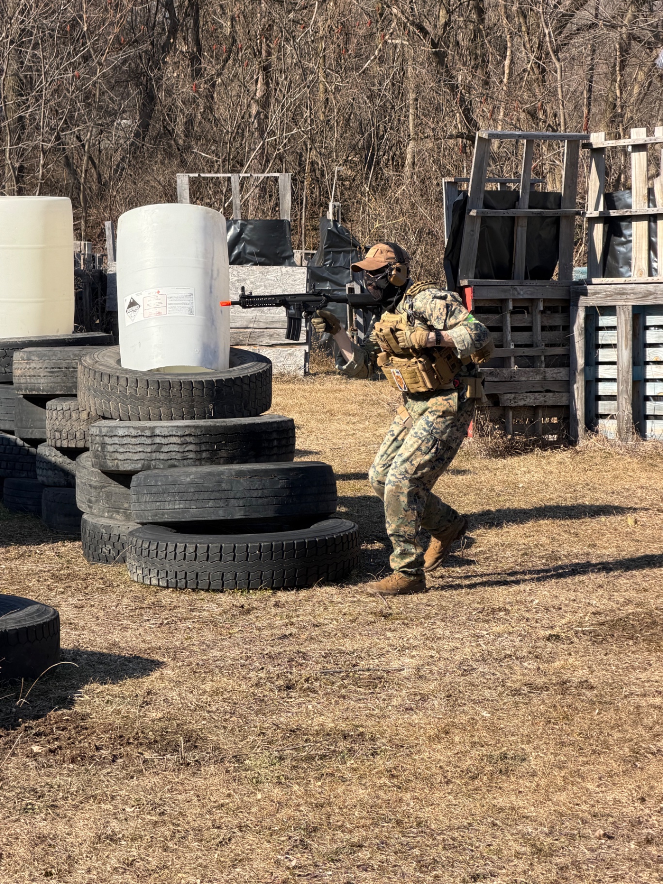 A player in camouflage aims an airsoft rifle from beside a tyre and pallet barrier