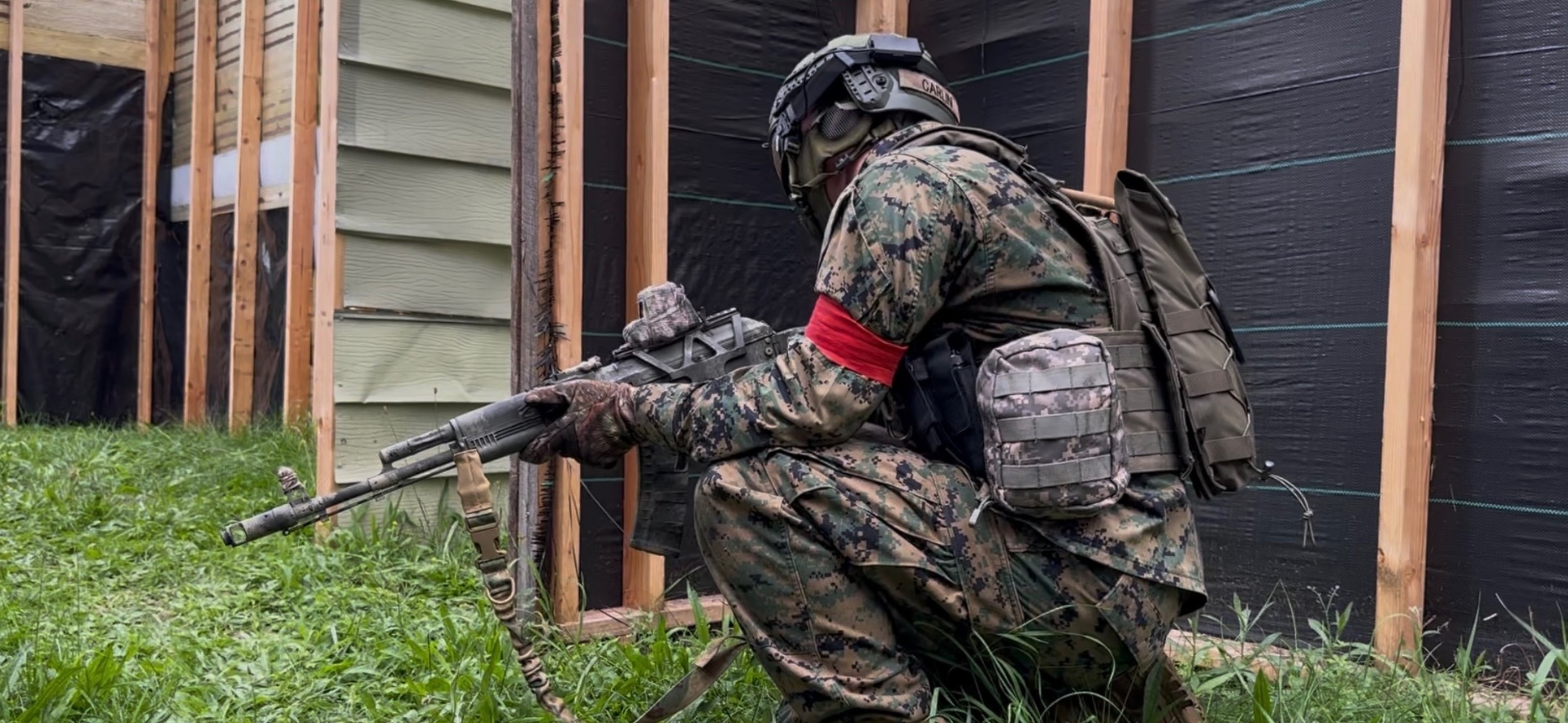 A player in digital camouflage, vest, backpack, red armband, helmet and goggles crouches in grass near a wooden frame covered with black sheeting
