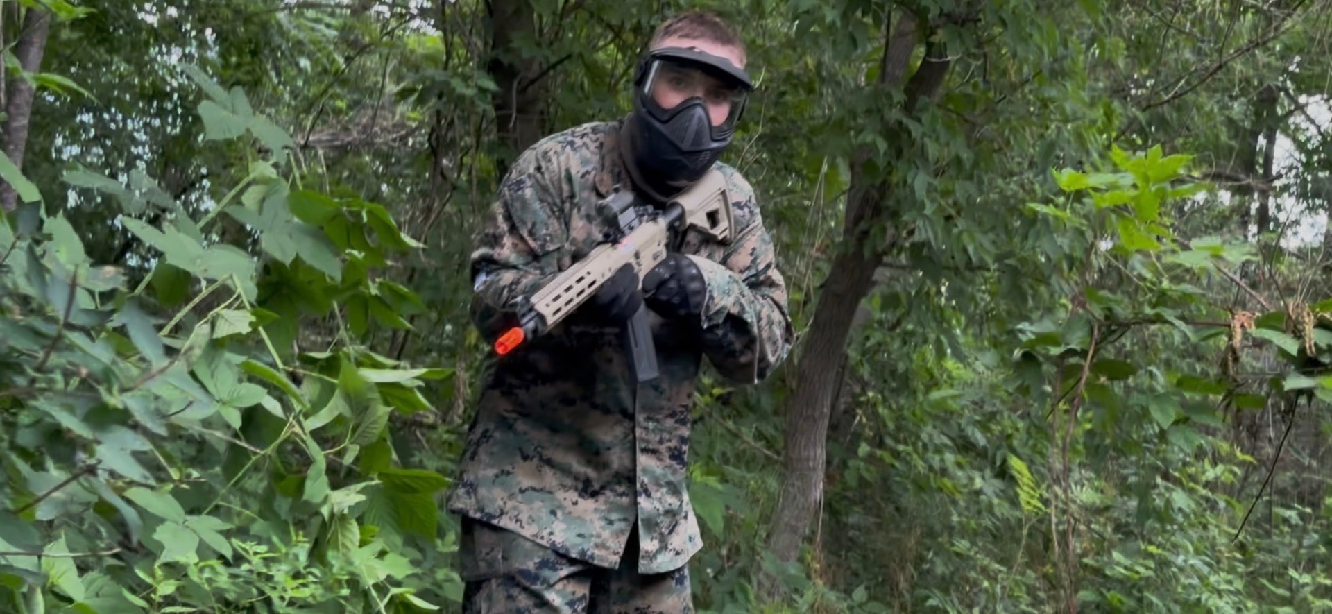 A player in digital woodland camouflage and a full-face protective mask aims an orange-tipped rifle among dense green trees