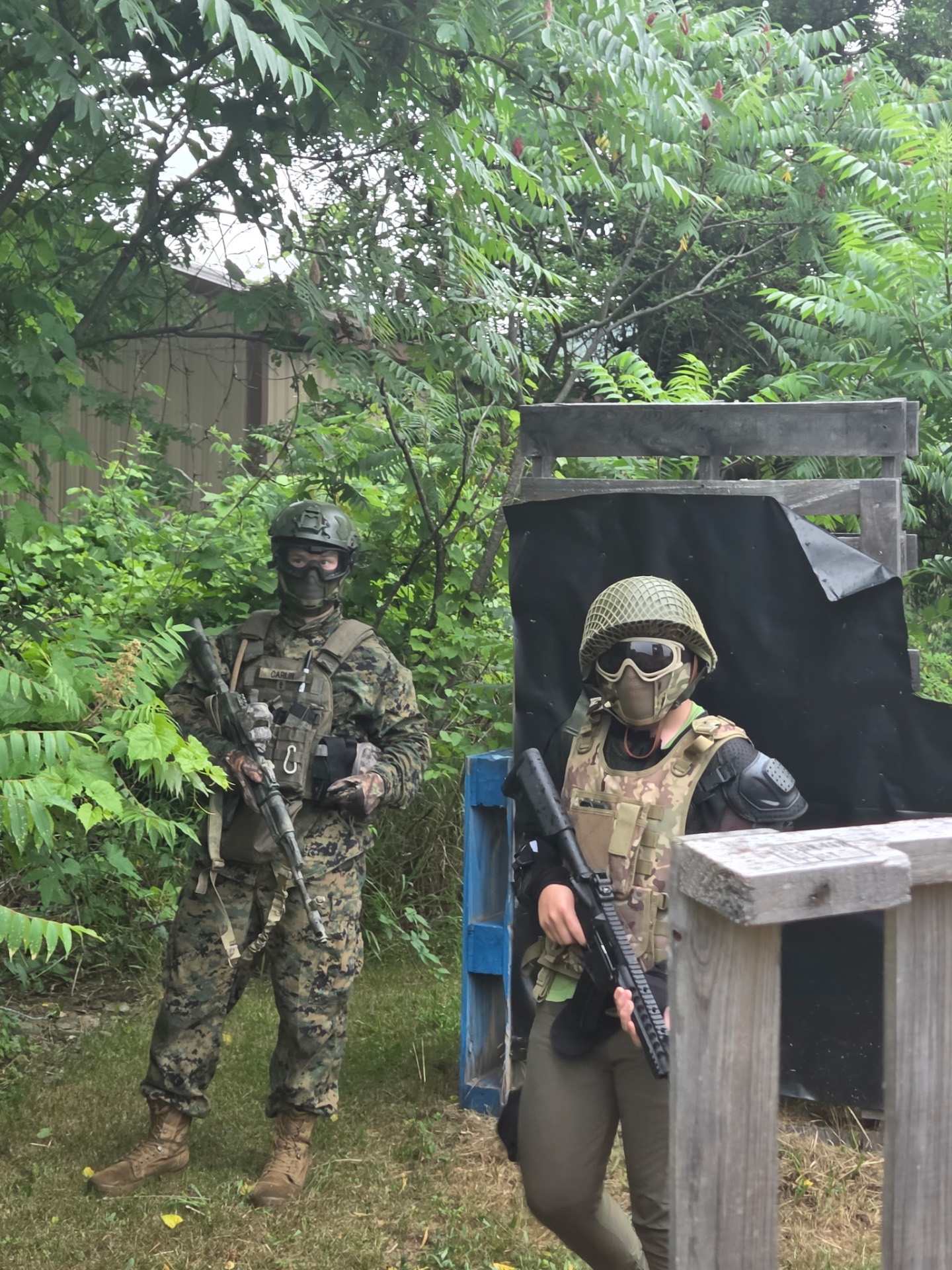 Two players in protective masks, helmets and tactical vests hold rifles beside wooden pallet cover surrounded by trees