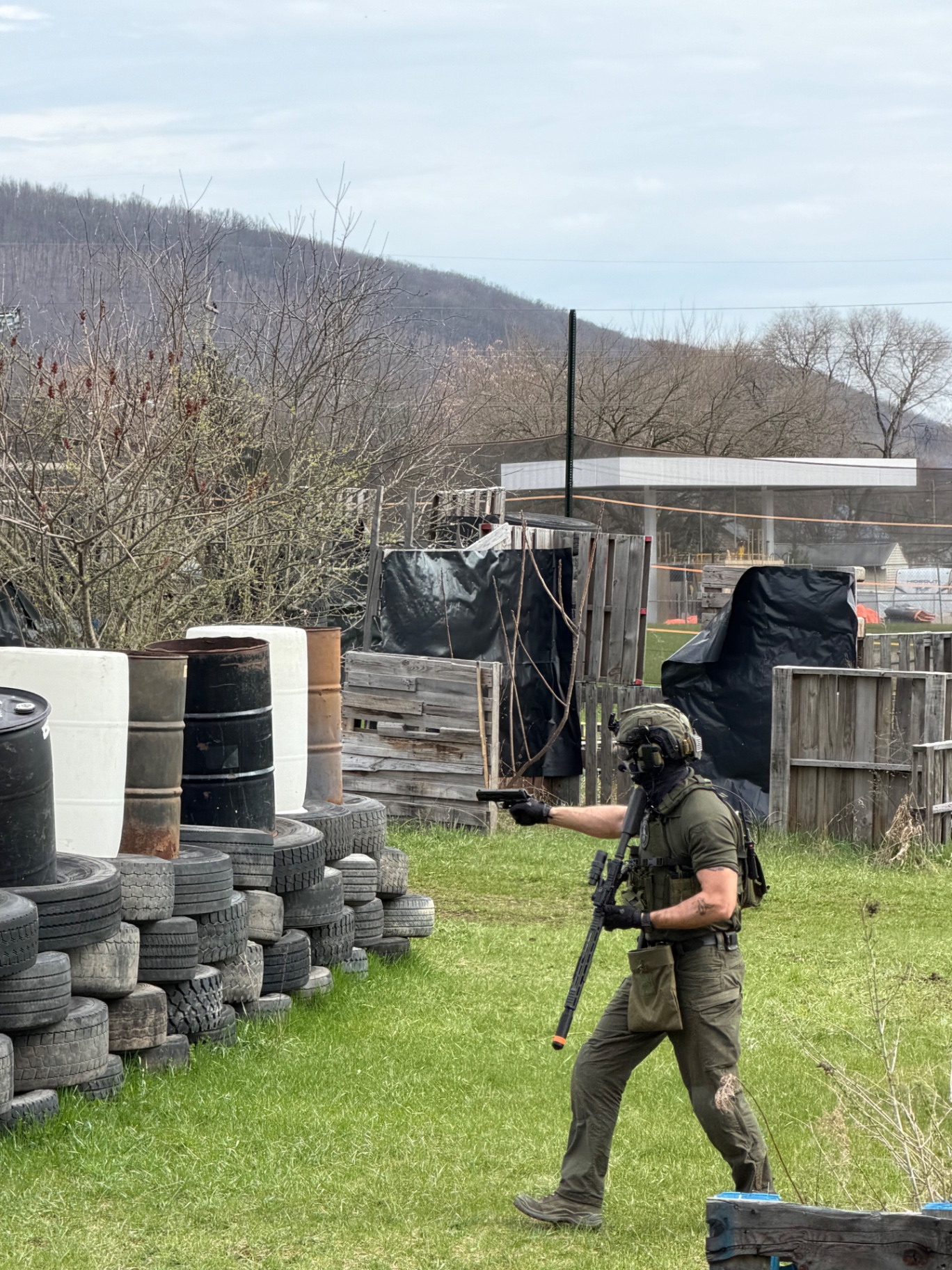 A player in an olive shirt, helmet and tactical vest aims a handgun across the Korhaven field while carrying a rifle, with tyre walls and drums around