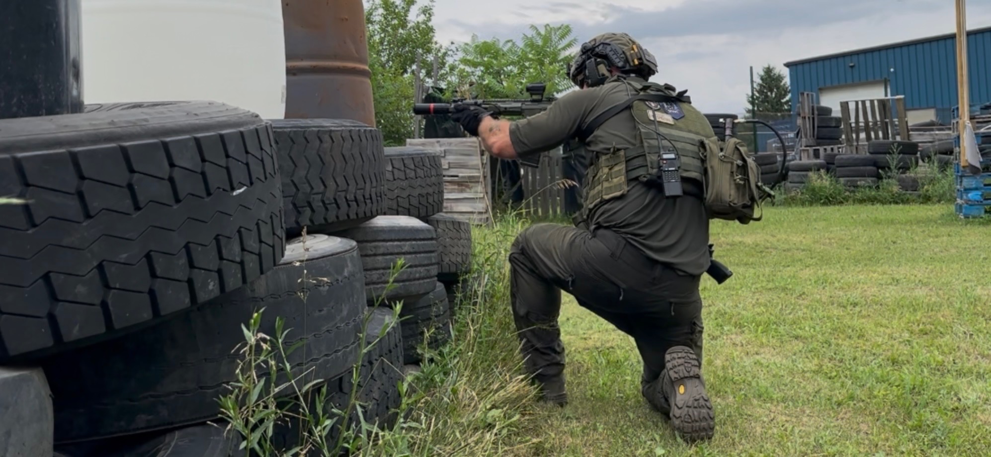 A player seen from behind in dark olive clothing kneels behind a barricade of stacked tyres and aims a rifle, with pallets and a building beyond