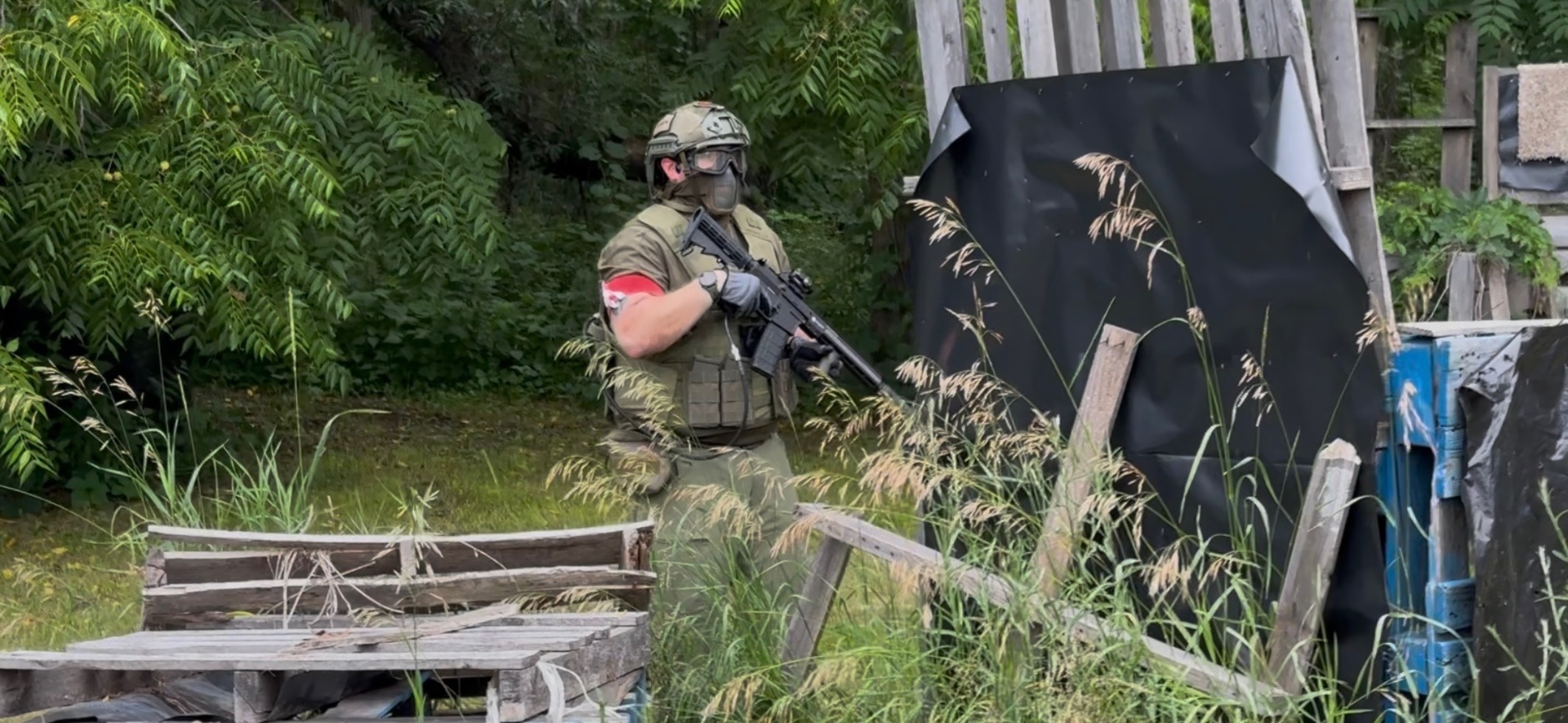 A player in an olive uniform, vest, helmet and lower-face mesh mask stands beside pallet barricades holding an AR-platform rifle