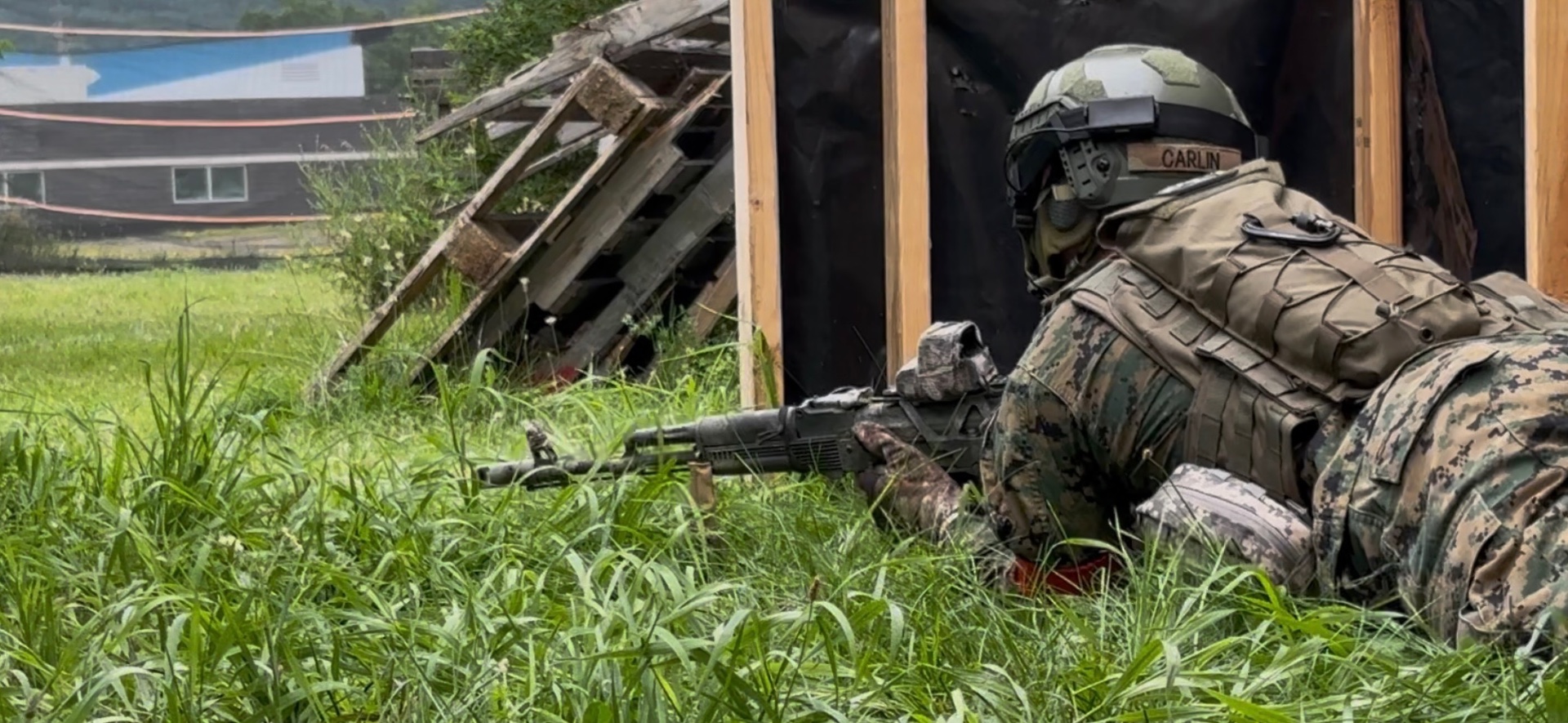 A player in MARPAT camouflage lies prone in tall grass aiming an AK-pattern rifle beside a plywood and black tarp barricade
