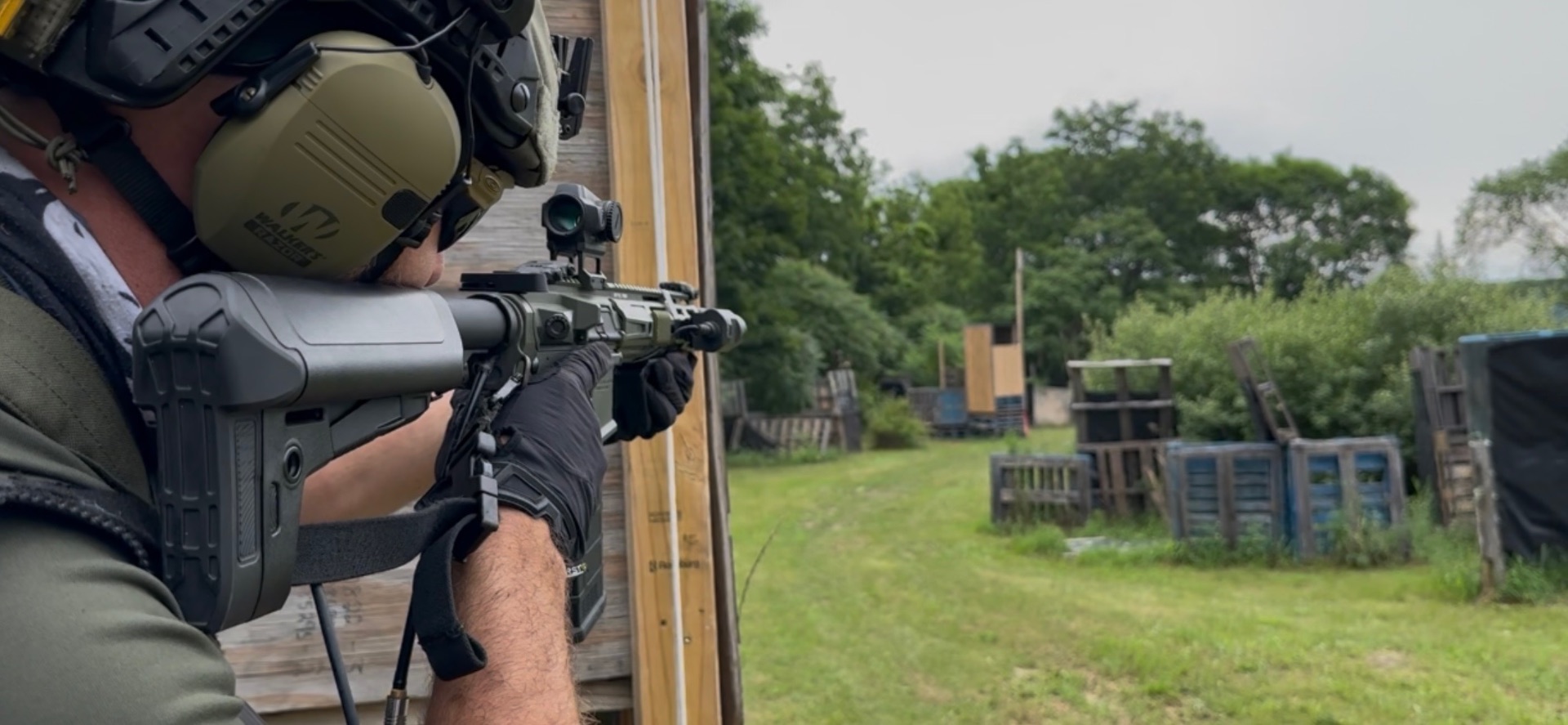 A player in a helmet, ear protection and gloves aims a rifle around a wooden barrier into a field of trees and pallet obstacles