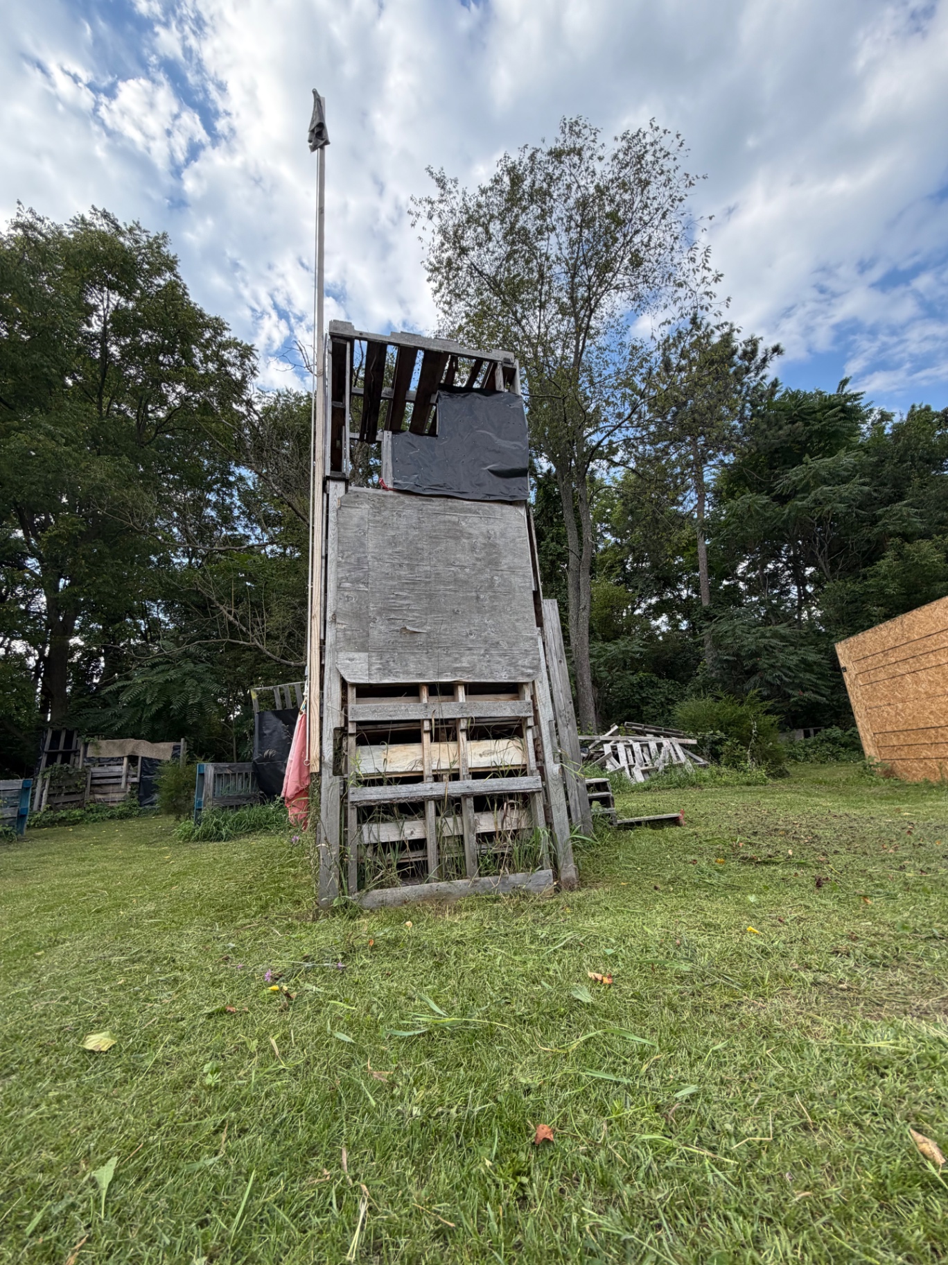 A tall tower built from wooden pallets and plywood stands on the grassy Korhaven field, surrounded by trees and makeshift barriers
