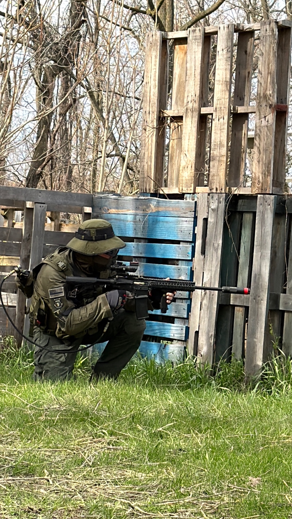 A player in an olive boonie hat and Airborne patch kneels on the grass aiming a scoped, orange-tipped airsoft rifle beside stacked wooden pallets