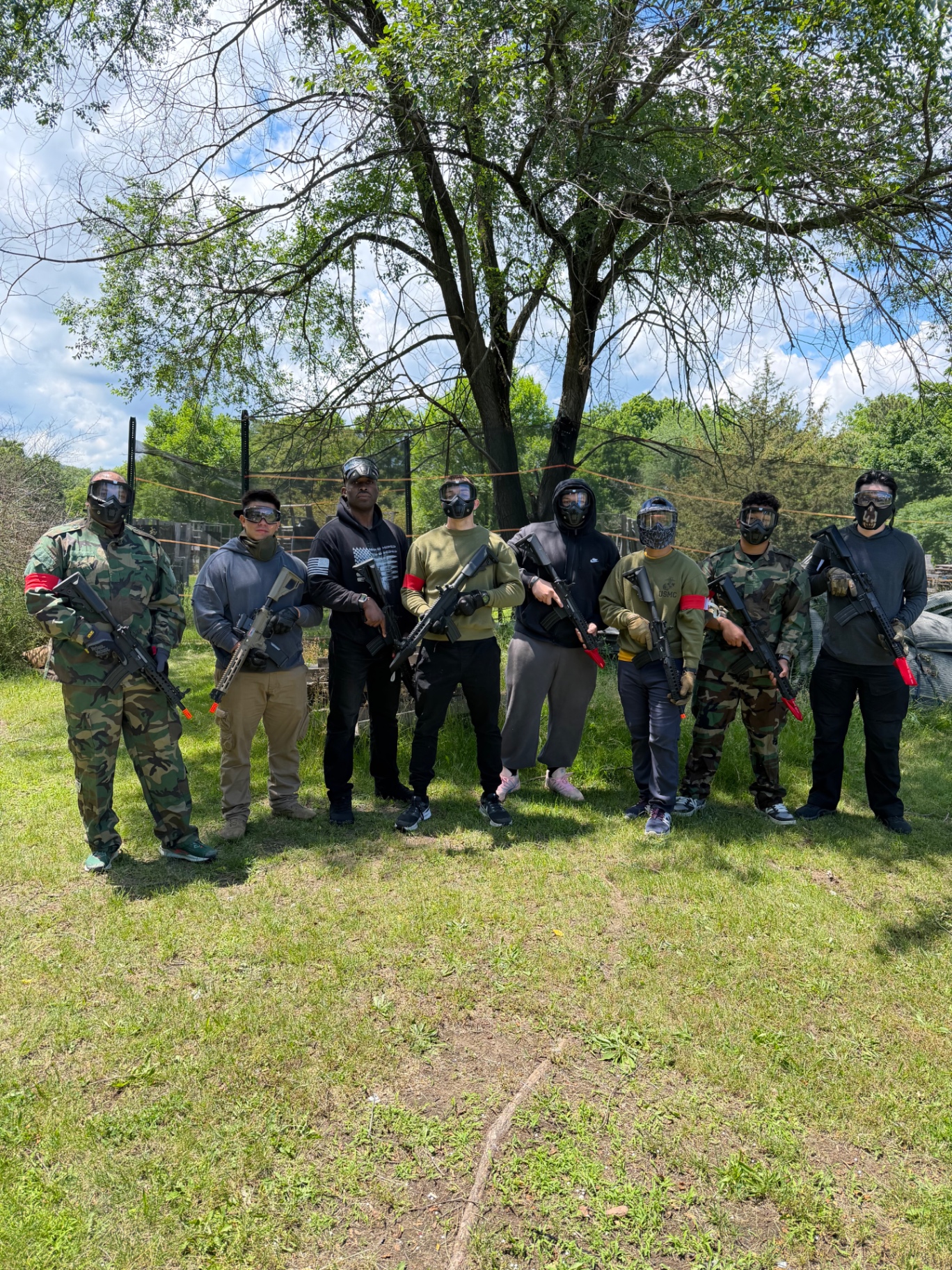 Eight players in face masks, red team armbands and mixed camouflage stand in a row on the grass holding rifles in front of a large tree and safety netting