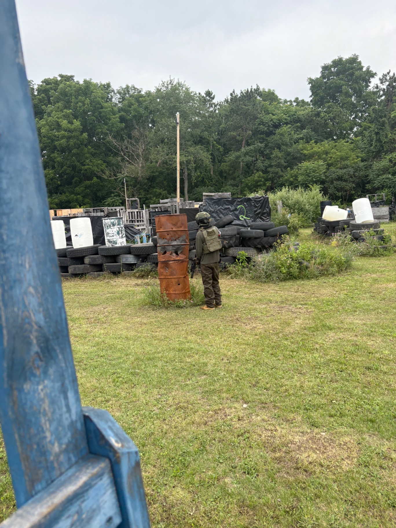 A player in olive uniform, helmet and plate carrier stands beside stacked rusty barrels near tyre, drum and pallet barricades, seen past a blue wooden post