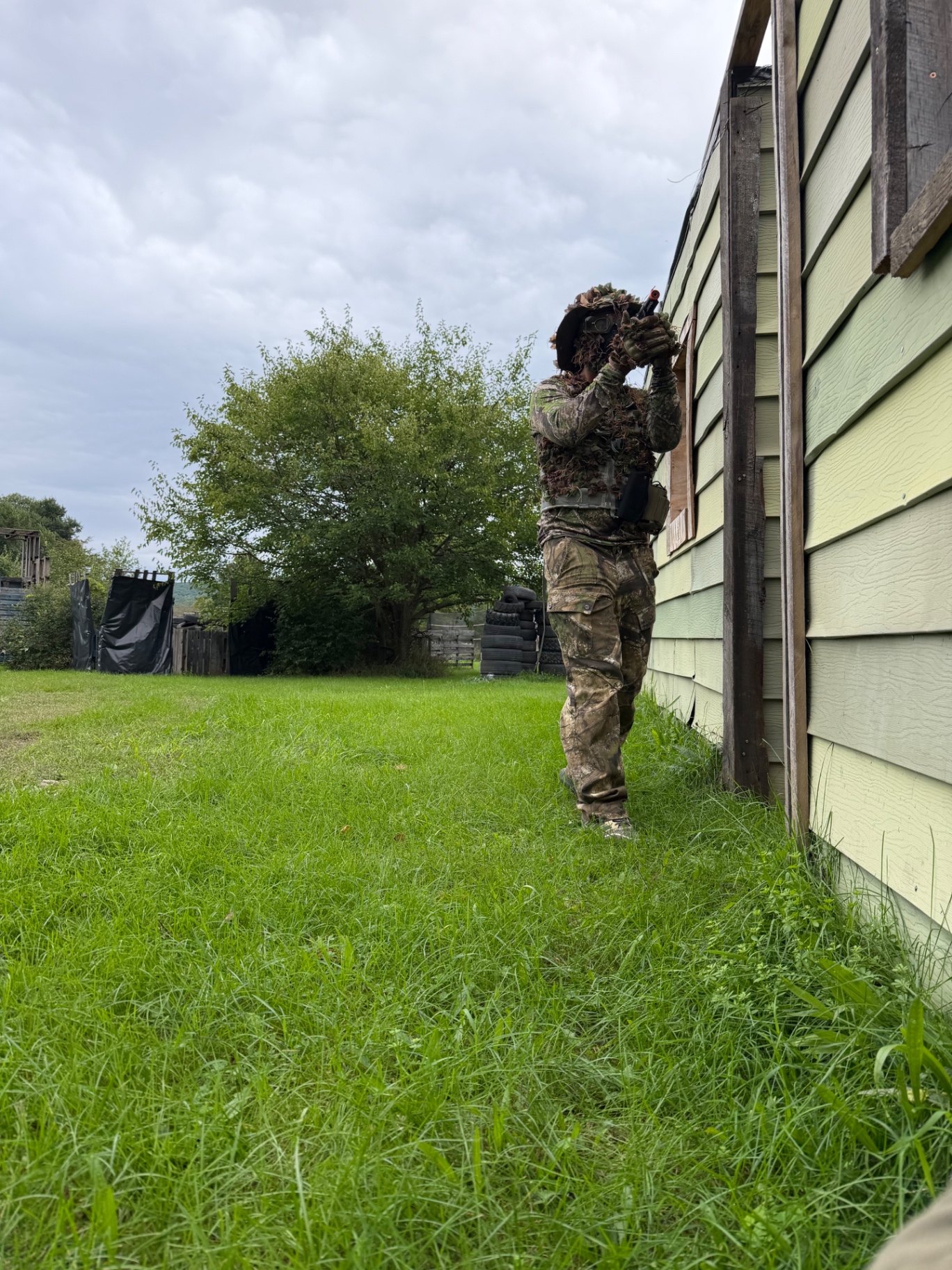 A player in ghillie camouflage aims an airsoft pistol while standing against the side of a light green wooden building