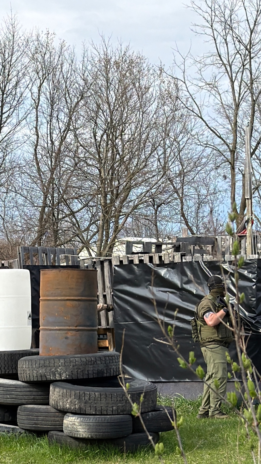 A player in olive tactical pants, a vest and a face wrap stands in profile beside pallet barricades, stacked tyres and barrels on the Korhaven field