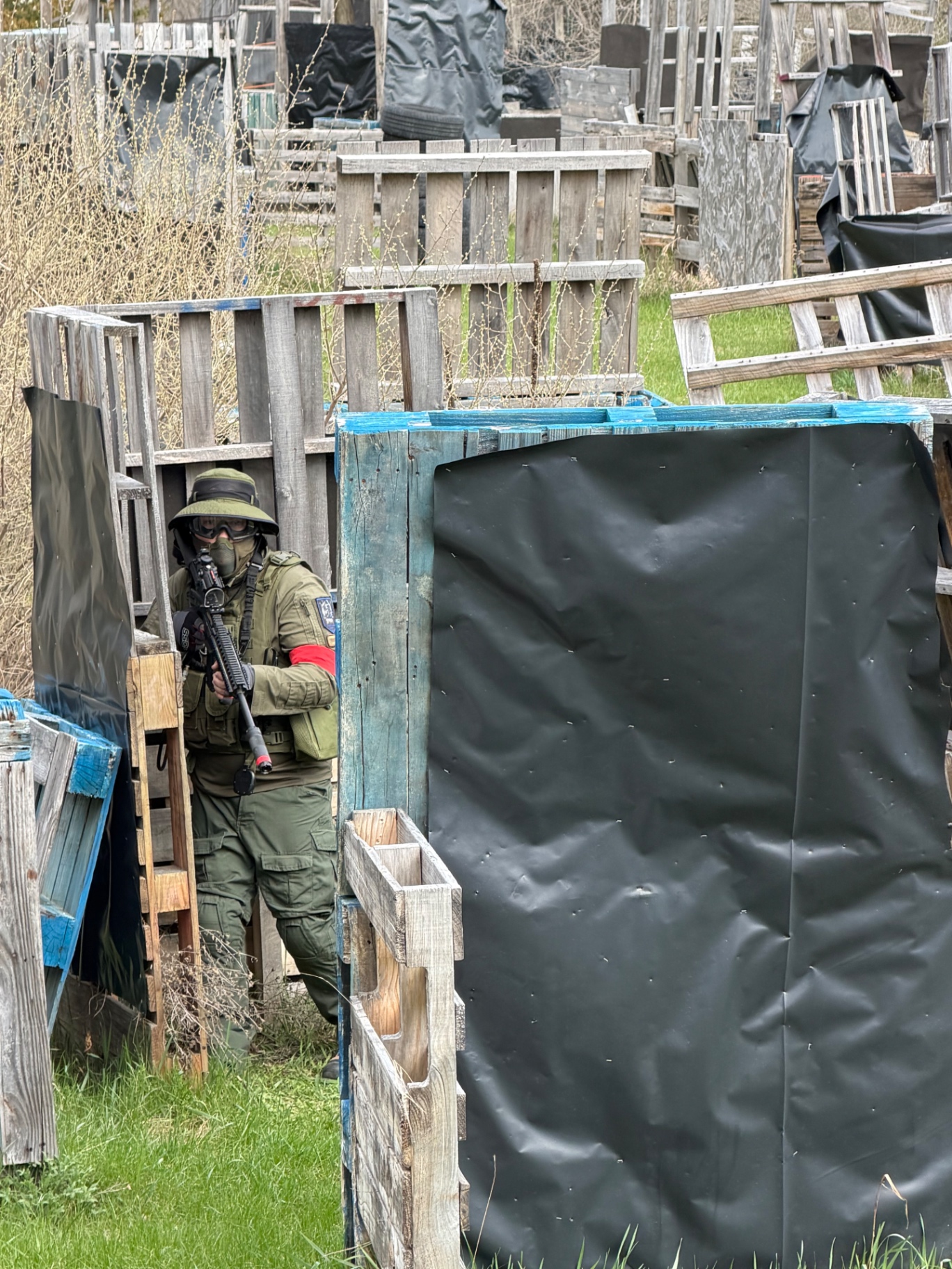 A player in an olive drab uniform, boonie hat and goggles aims an orange-tipped airsoft rifle from behind pallet barricades covered in black sheeting