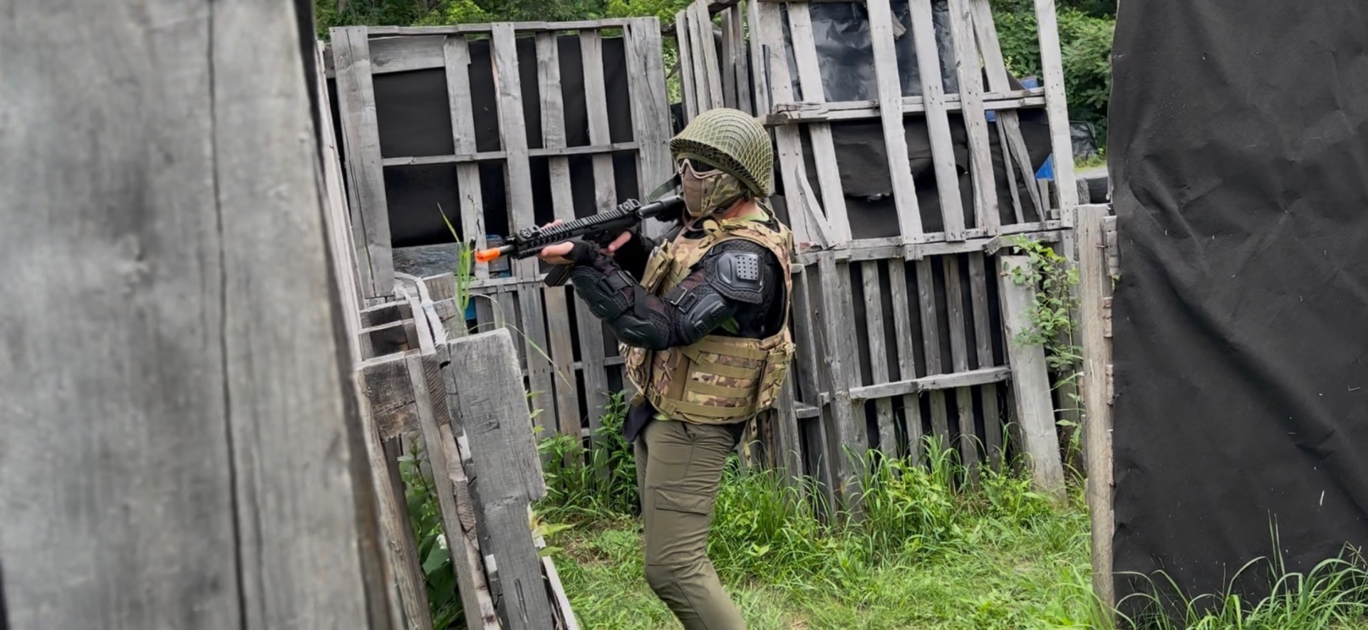 A player in a netted helmet, tactical vest and elbow pads aims an airsoft rifle behind wooden pallet barriers in a grassy field