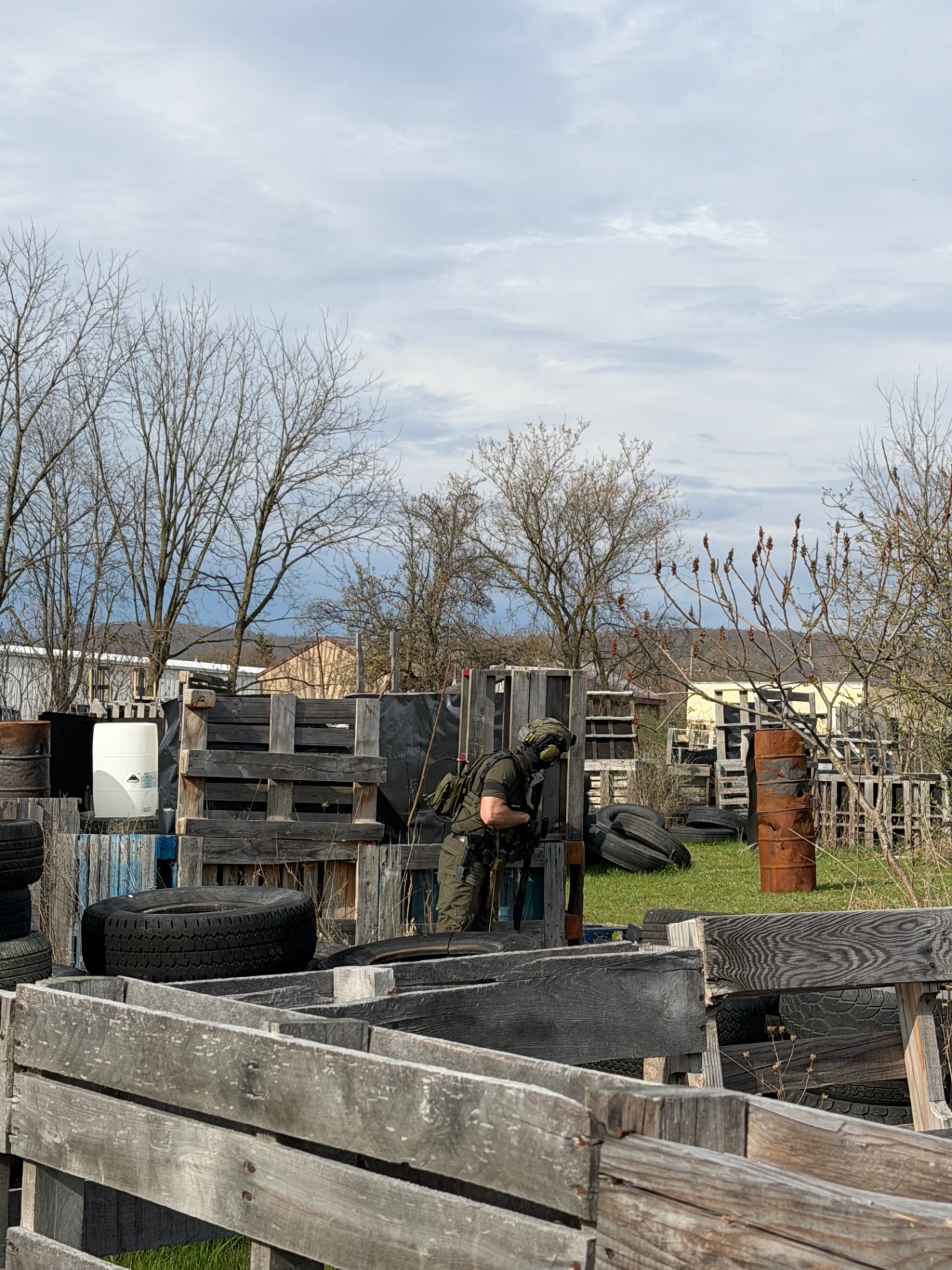A wide view of a player in green gear moving behind pallet barricades among stacked tyres and metal barrels on the Korhaven field