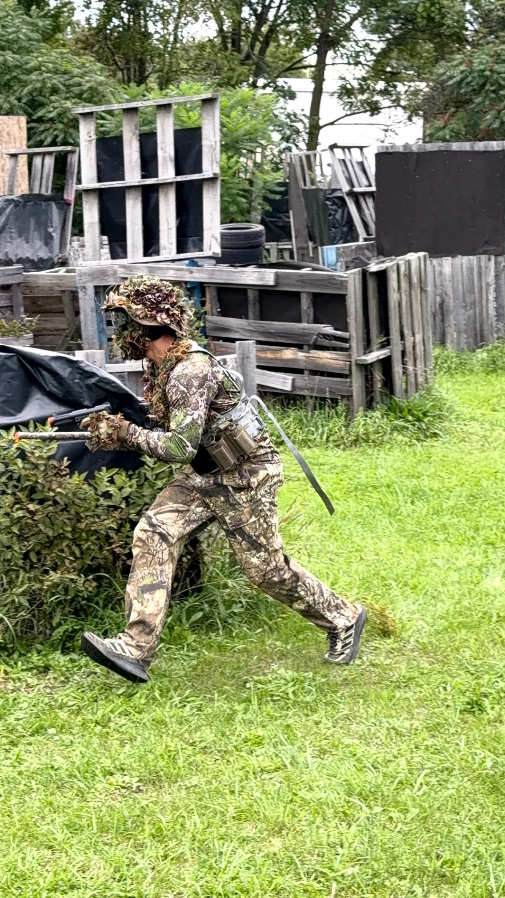 A player in camouflage and a ghillie-style hat runs across the grass past pallet barricades carrying an airsoft gun