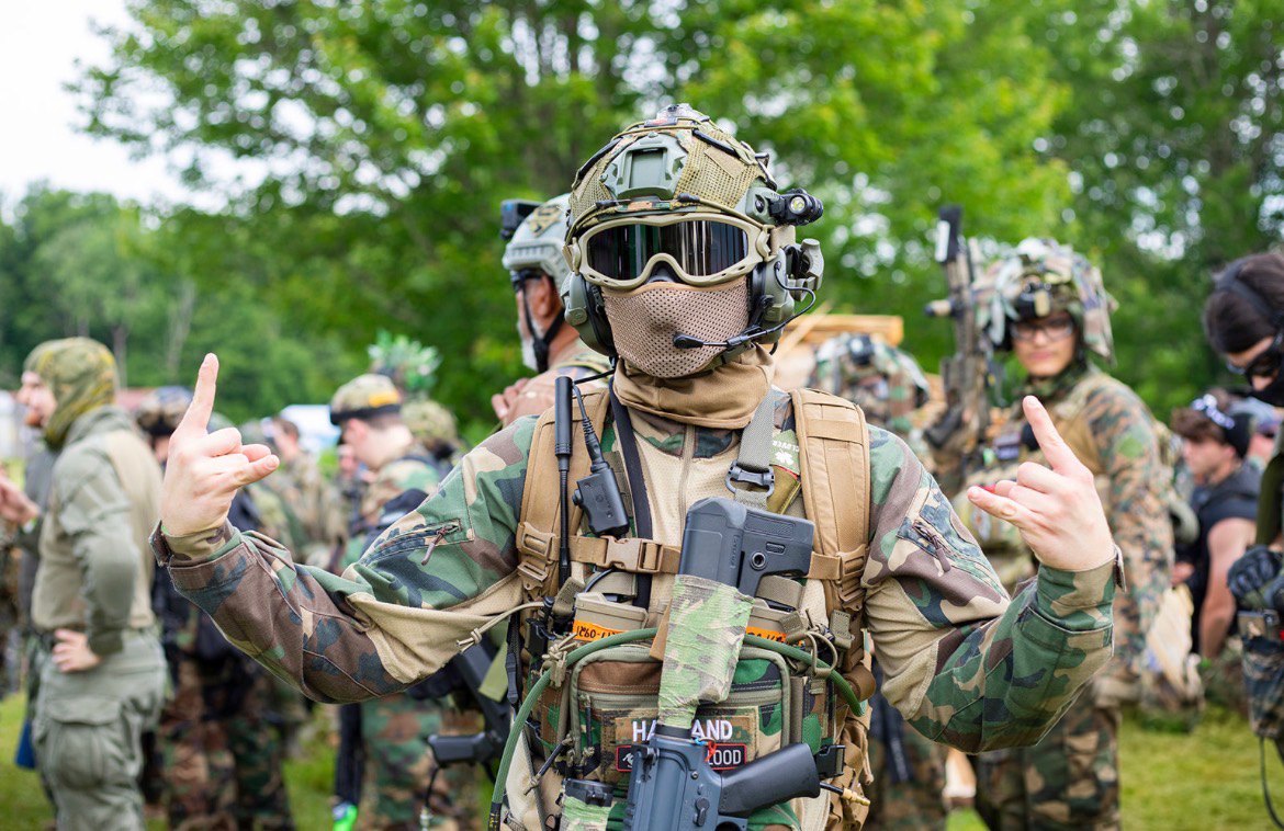 A player in a mesh mask, goggles, helmet and chest rig faces the camera flashing a hand sign while other players and trees stand behind