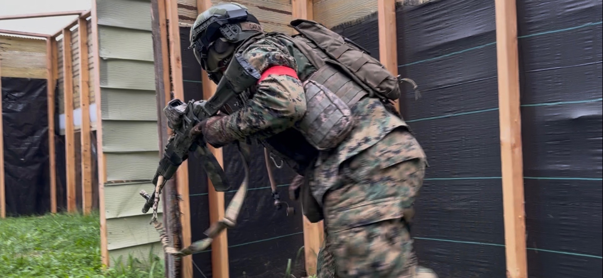 A player in digital camouflage and a red armband advances past wooden and black tarp barricades carrying an AK-pattern rifle