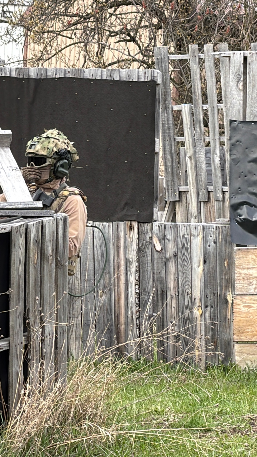 A player wearing a camouflage-covered helmet, ear protection, goggles and a lower face mask stands behind wooden pallet barriers on the Korhaven field