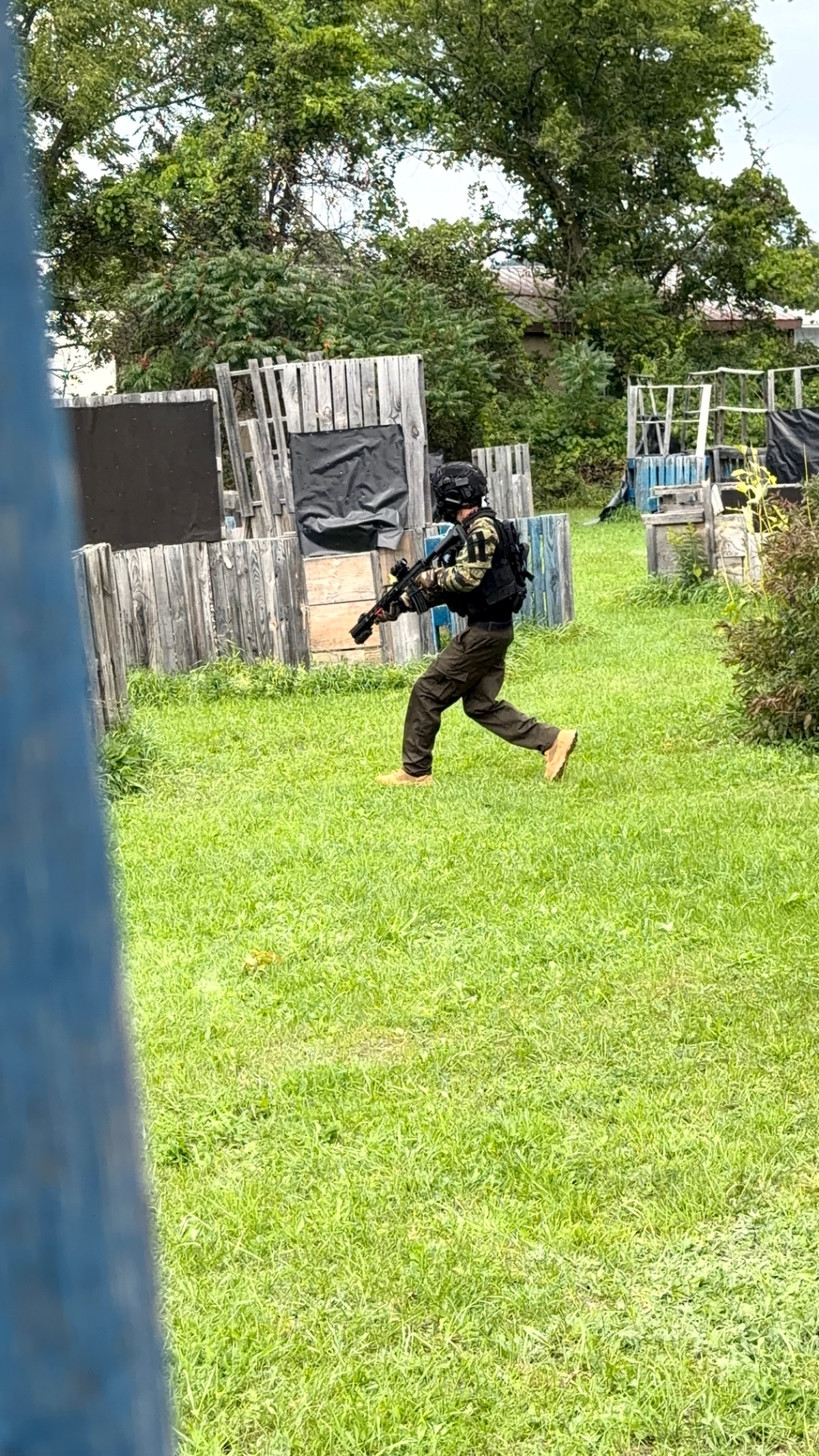 A player in tactical gear and camo sleeves sprints across open grass carrying a tactical rifle, with pallet structures and trees around