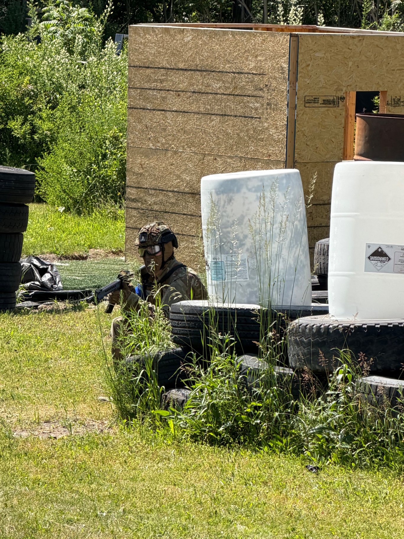 A player in camouflage, helmet and goggles aims a rifle from behind stacked tyres, white plastic drums and a plywood structure