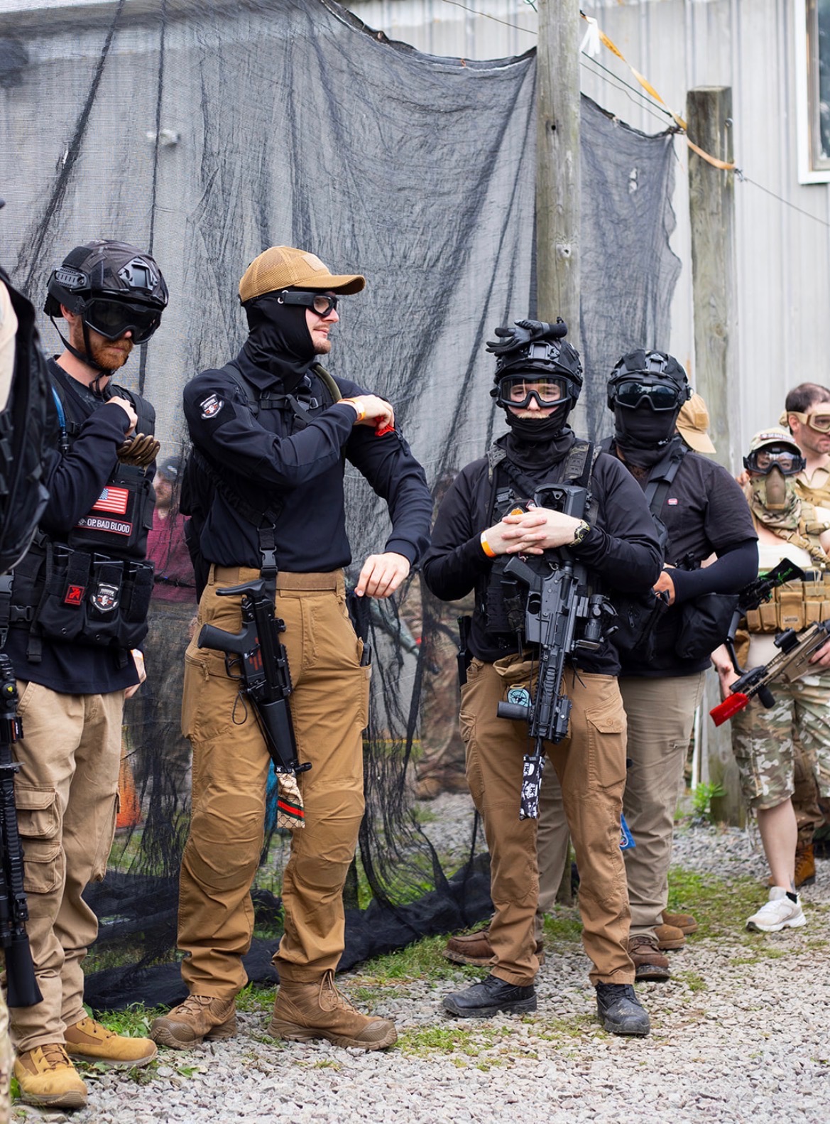 Players in goggles, helmets, plate carriers and tan cargo pants stand in a gravel staging area beside safety netting, one holding an airsoft rifle