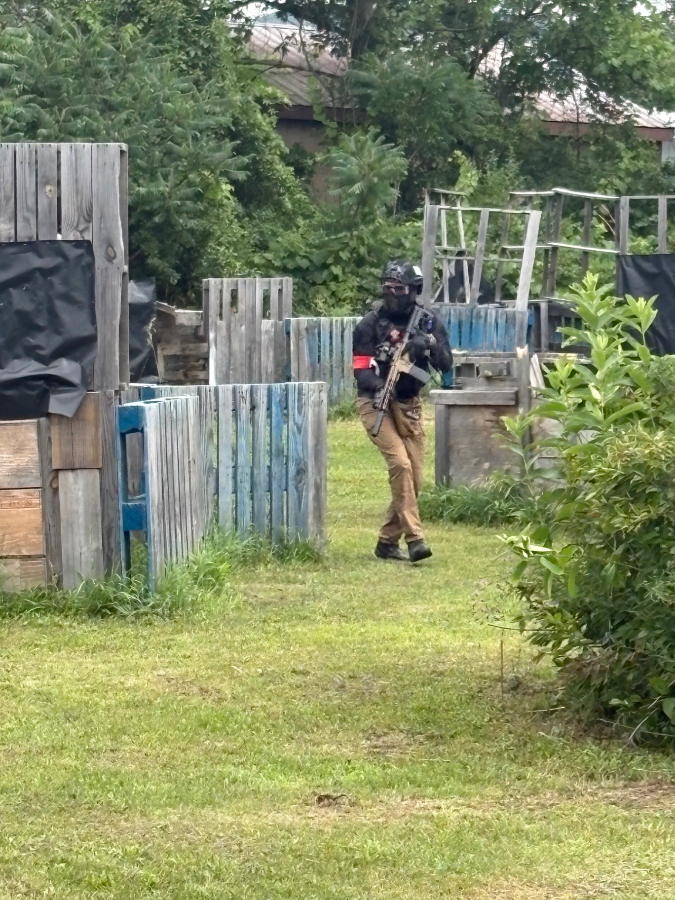 A player in a helmet, full face mask, tactical vest, red armband and tan pants carries a rifle along wooden pallet barriers on a grassy field