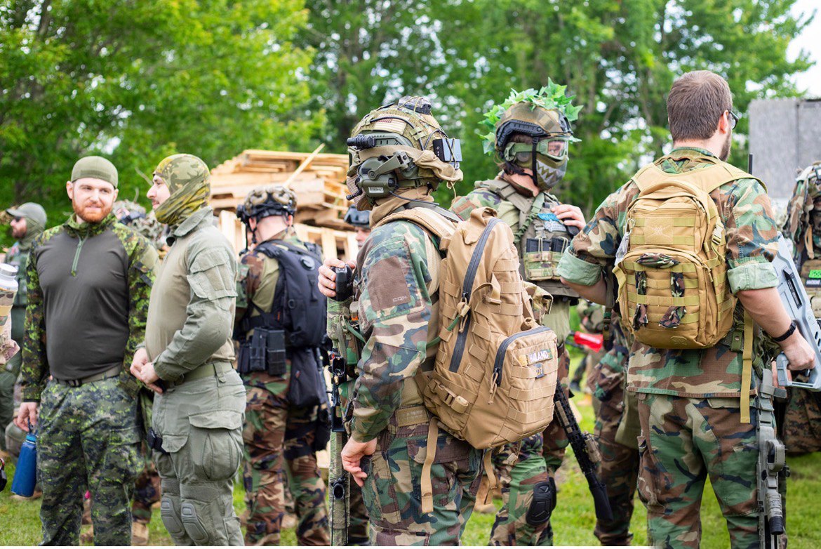 Players in various camouflage uniforms, helmets and backpacks stand and talk together in a grassy field bordered by trees