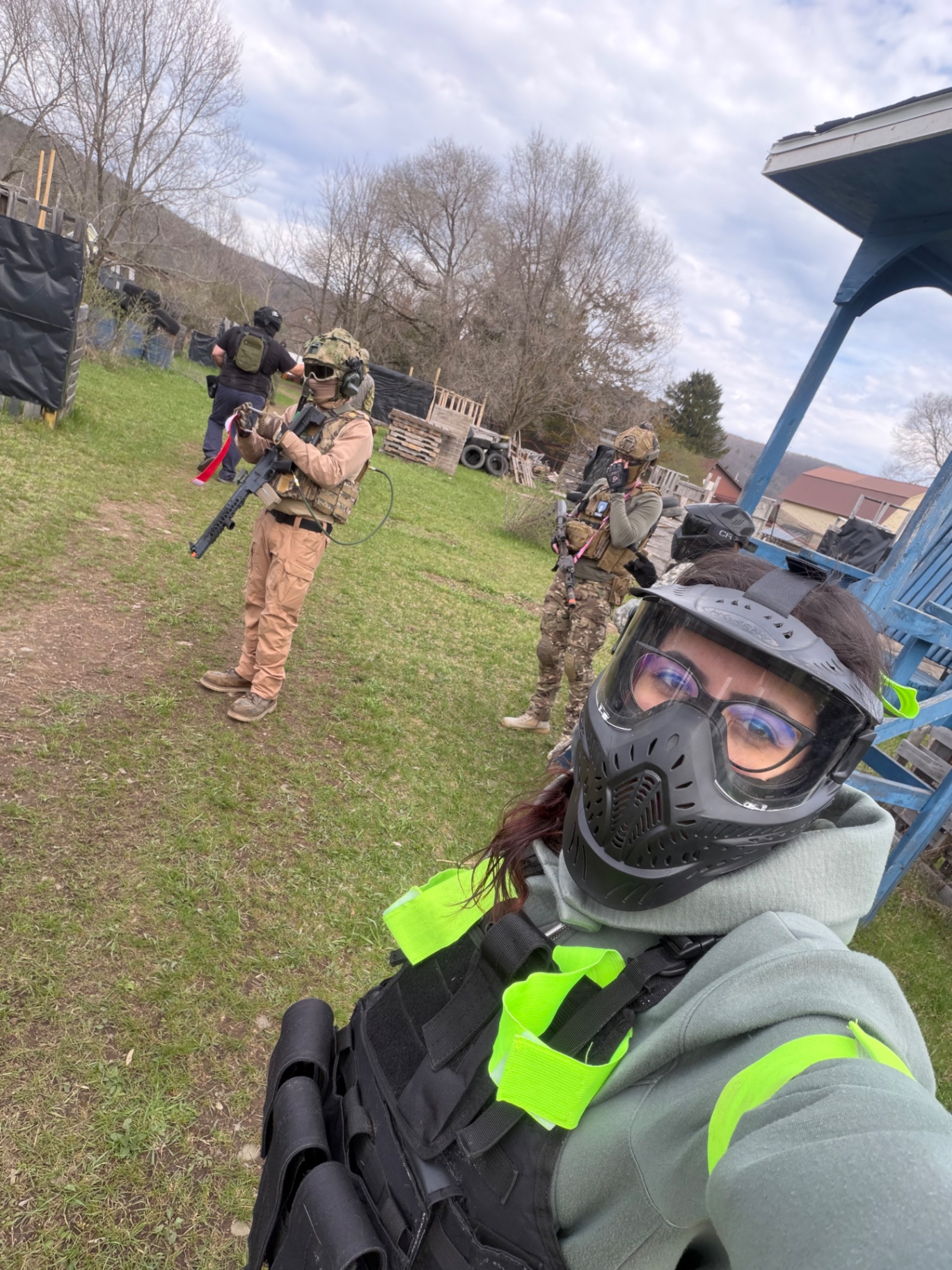 A player in a full-face protective mask, tactical vest and hi-vis straps takes a selfie while other players with airsoft rifles stand ready on the Korhaven field