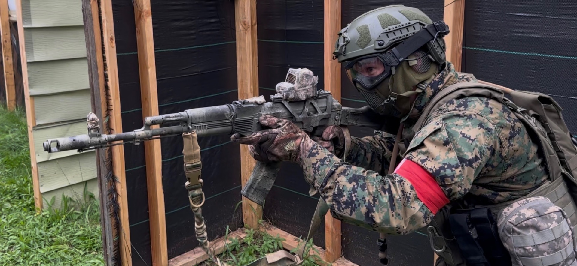 A player in full tactical gear with a red armband aims a painted AK-style rifle from beside a plywood and tarp barricade