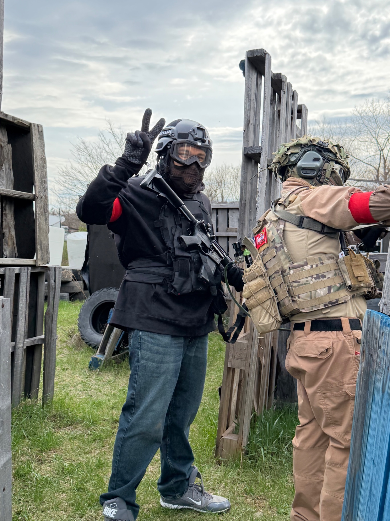 Two players in helmets, goggles and red team armbands stand beside wooden pallet barriers on the Korhaven field, one holding an airsoft rifle and flashing a peace sign
