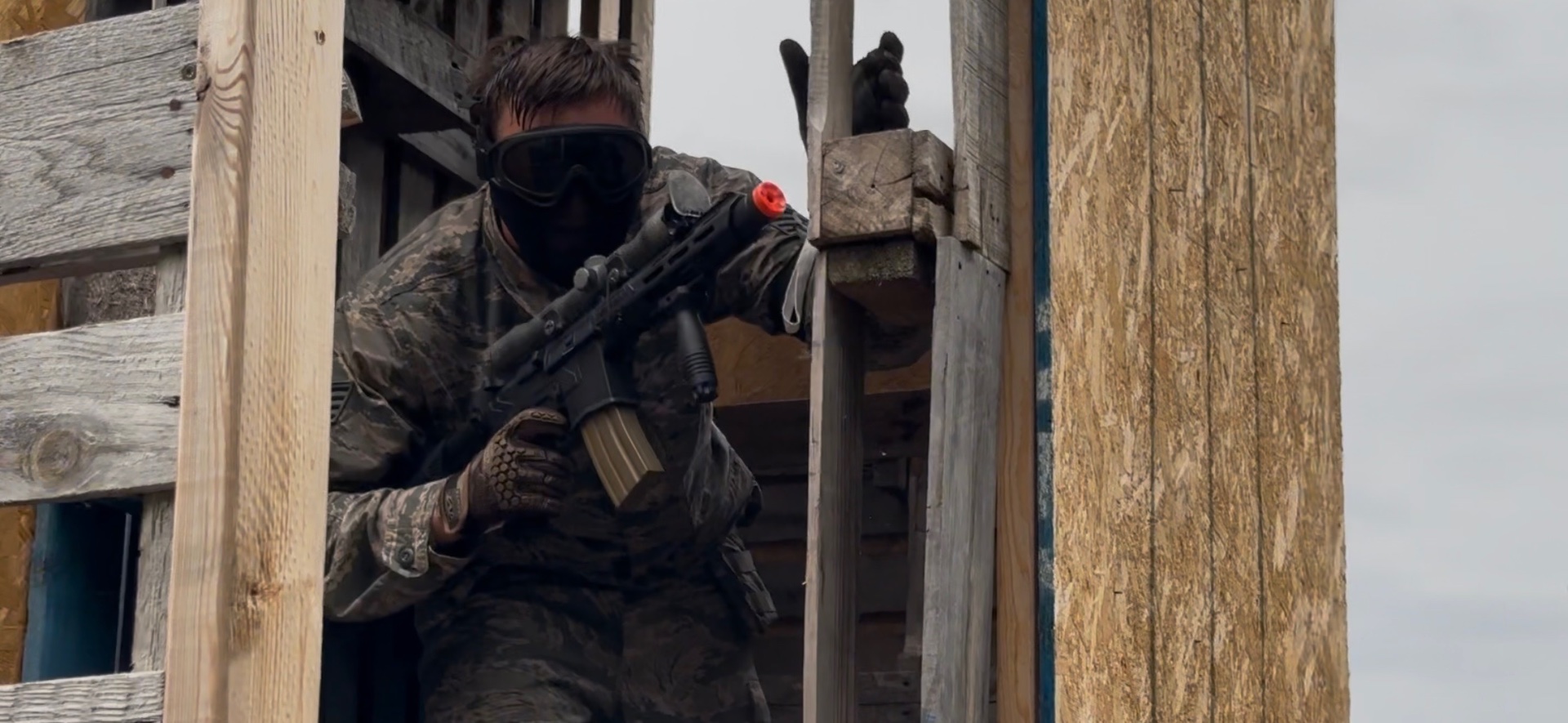 A player in camouflage fatigues, goggles and a face mask holds an orange-tipped rifle behind a wooden pallet and plywood structure