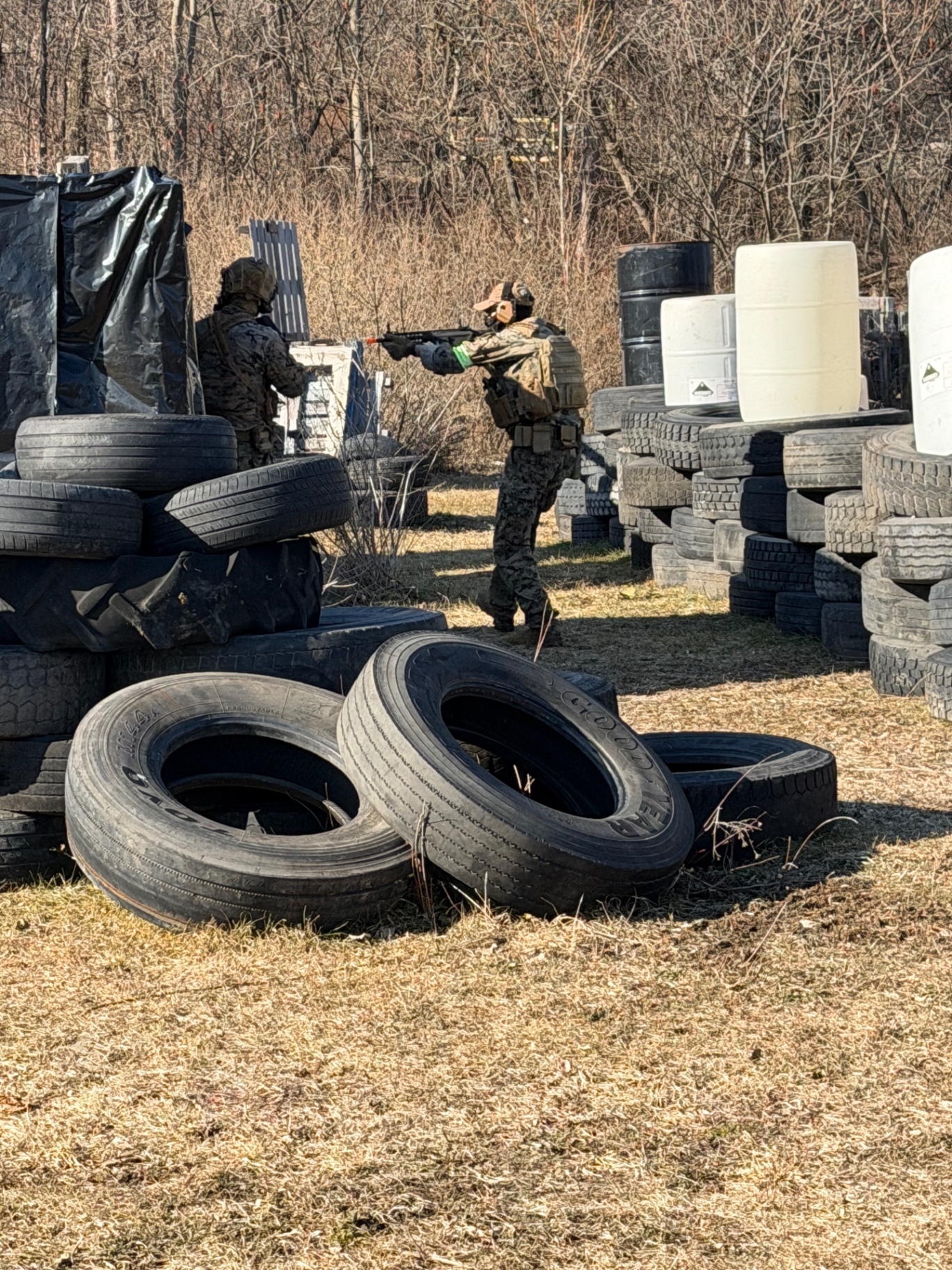 Two players in camouflage and tactical gear holding airsoft rifles among stacks of tyres, plastic drums and wooden pallets on the Korhaven field