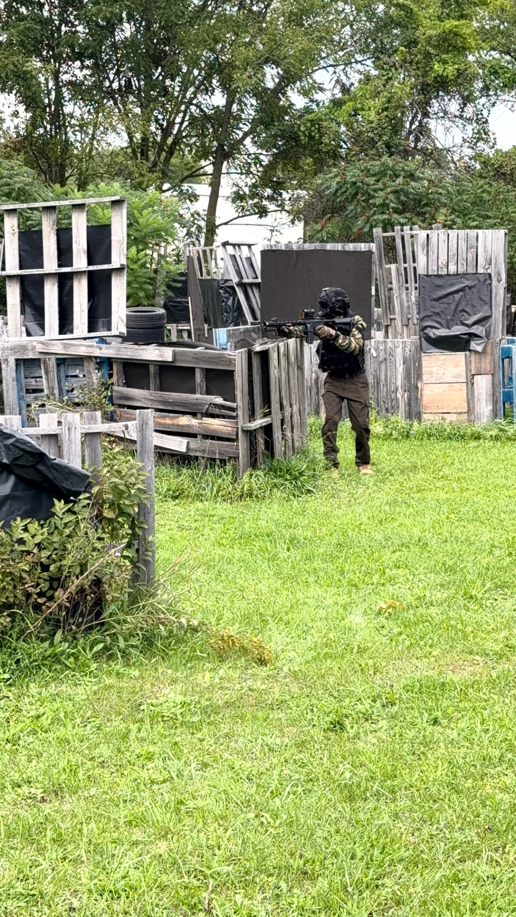A player in camouflage, dark tactical vest, helmet and face protection aims a tactical rifle among pallet and black tarp barriers